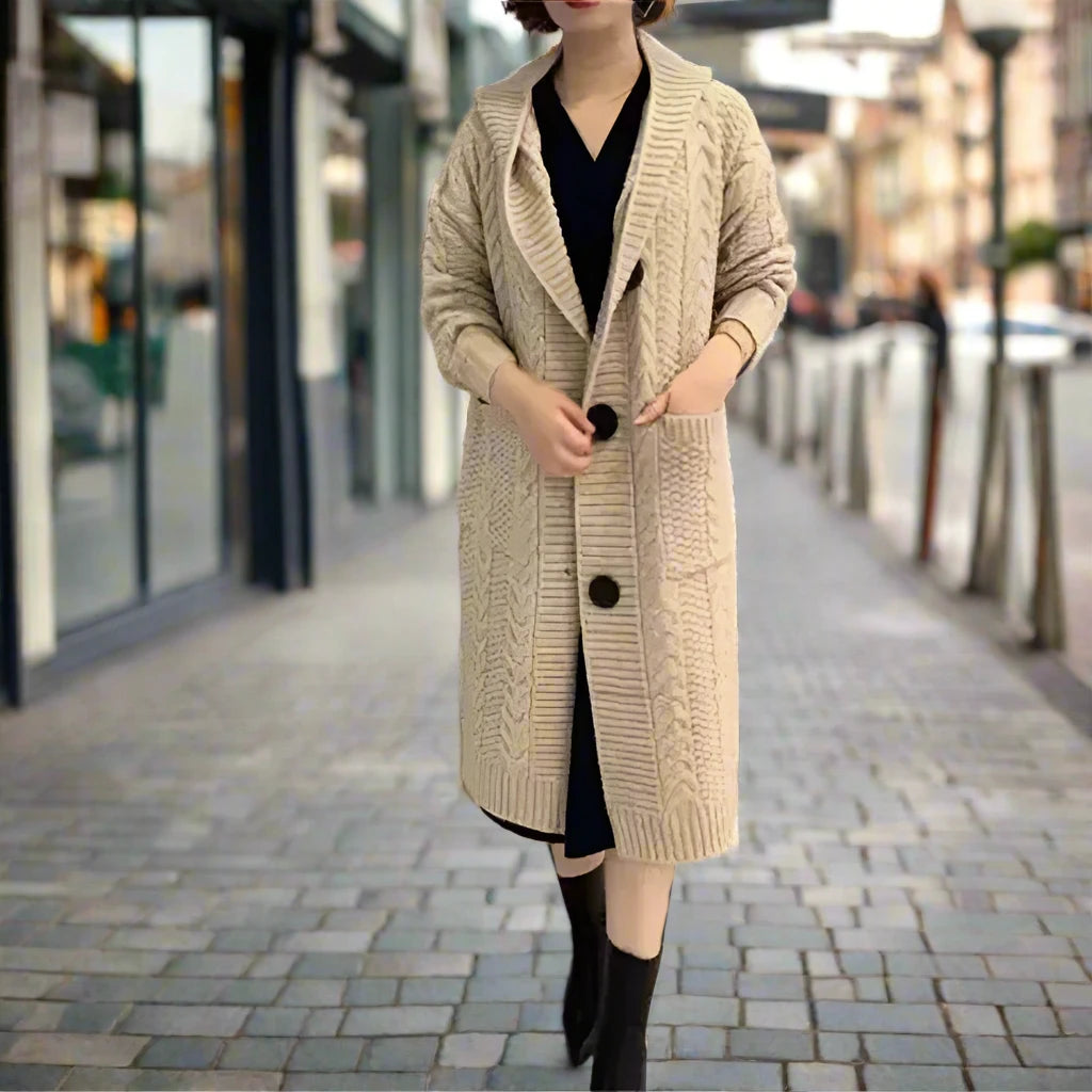 A person stands on a city street wearing a long apricot jacquard twist cardigan with large buttons, a black dress underneath, and black boots. The background is blurred, suggesting an urban setting with buildings and street elements.