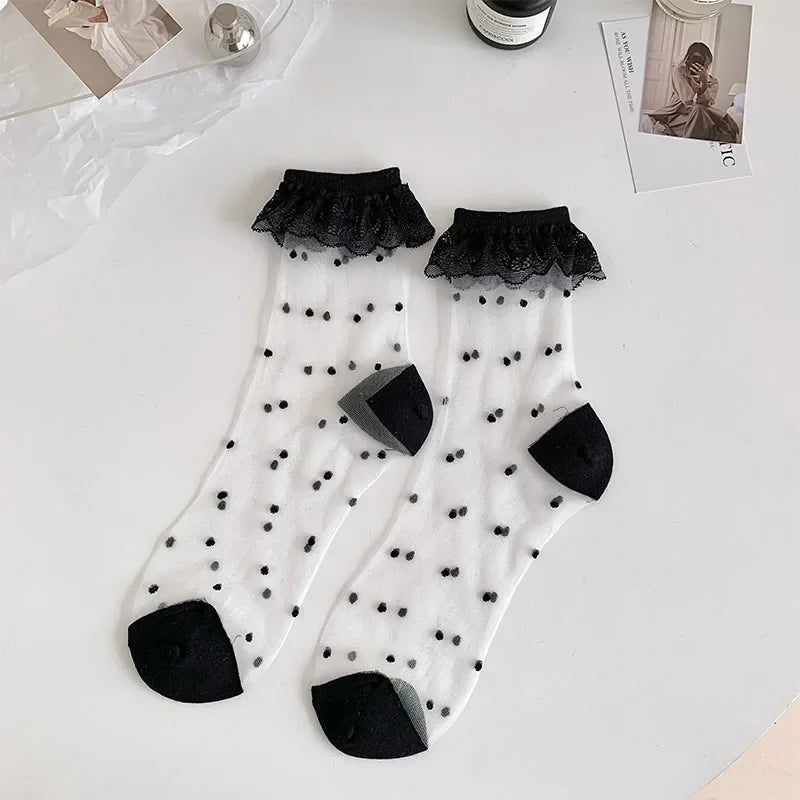 A pair of semi-transparent socks with a black dot pattern and black lace frills at the top are laid out on a white surface. The socks have black reinforced toes and heels. Surrounding the socks are a few items, including a silver spherical object, a bottle, and some photographs.