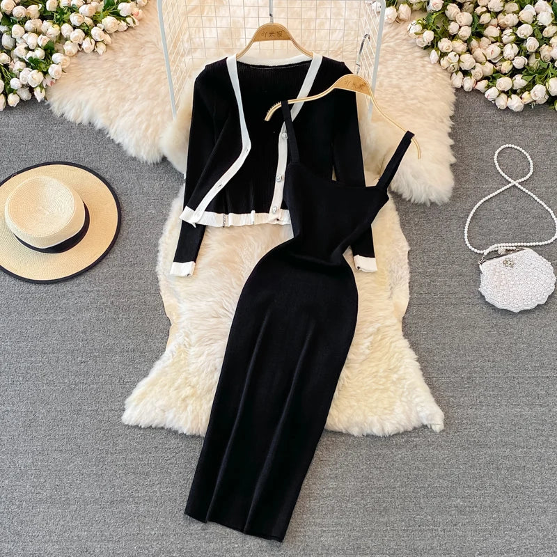 A black women’s outfit displayed on a white furry surface. The outfit includes a sleeveless dress with a draped neckline and a matching cardigan with white trim. Accessories include a white handbag and a beige hat on the left side, with white flowers and part of a wicker chair in the background.