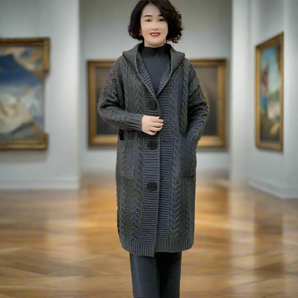 An individual with a blurred face stands in the center of an art gallery, wearing a long, Black grey cable knit cardigan over a black outfit. The person is holding one hand over the other in front of them. In the background, classic framed paintings are displayed on the gallery walls, creating an atmosphere of cultural and artistic significance.