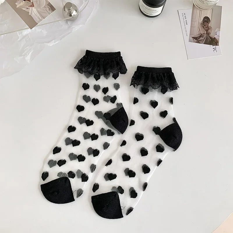 A pair of semi-transparent socks with a black heart pattern and black lace frills at the top are laid out on a white surface. The socks have reinforced black toes and heels. Surrounding the socks are a few items, including a silver spherical object, a bottle, and some photographs.