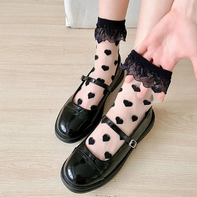 A close-up of a person's feet wearing sheer socks with black heart patterns and black lace trim at the top. The person is also wearing black patent leather Mary Jane shoes with a buckle strap. The background is a light-colored wooden floor. The combination of the sheer socks with heart patterns and the shiny black shoes creates a stylish and visually appealing look.