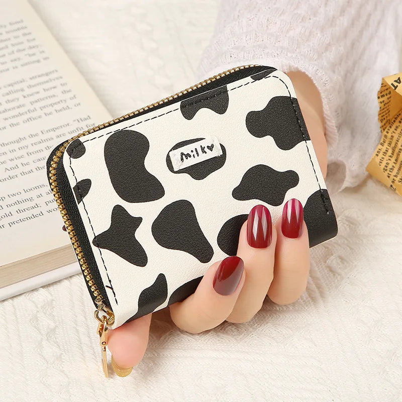 A showcasing of the Black and White Cow Print Card Bag being held in a hand to show the size comparison. It has a little tag on the front that says milk.