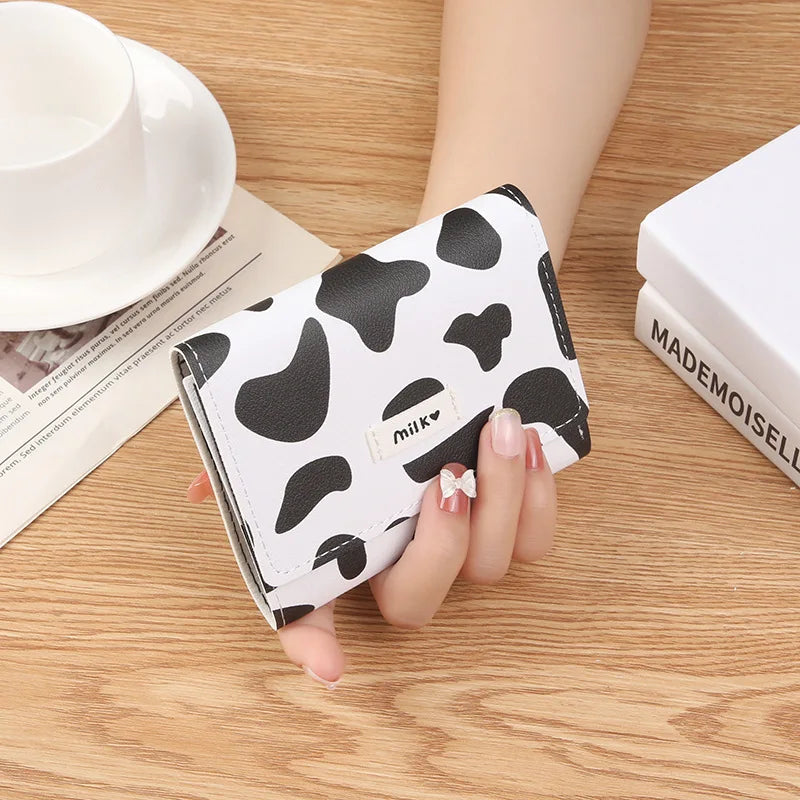 A showcase of the Black and White Cow Print Wallet with a little tag on the front that says milk being held in a hand to give a size comparison.