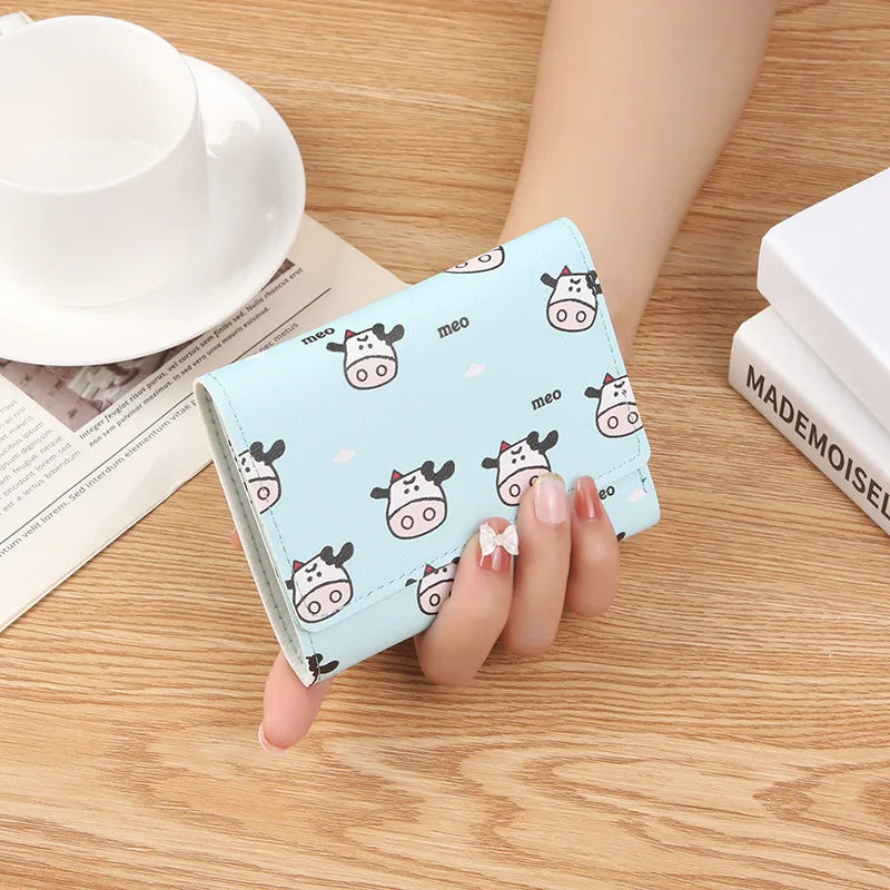 A showcasing of the Blue Cow and Cloud Meo Wallet being held in a hand to show the size comparison. This print is some cute cows with small white clouds and the word meo on top of a blue colored wallet.