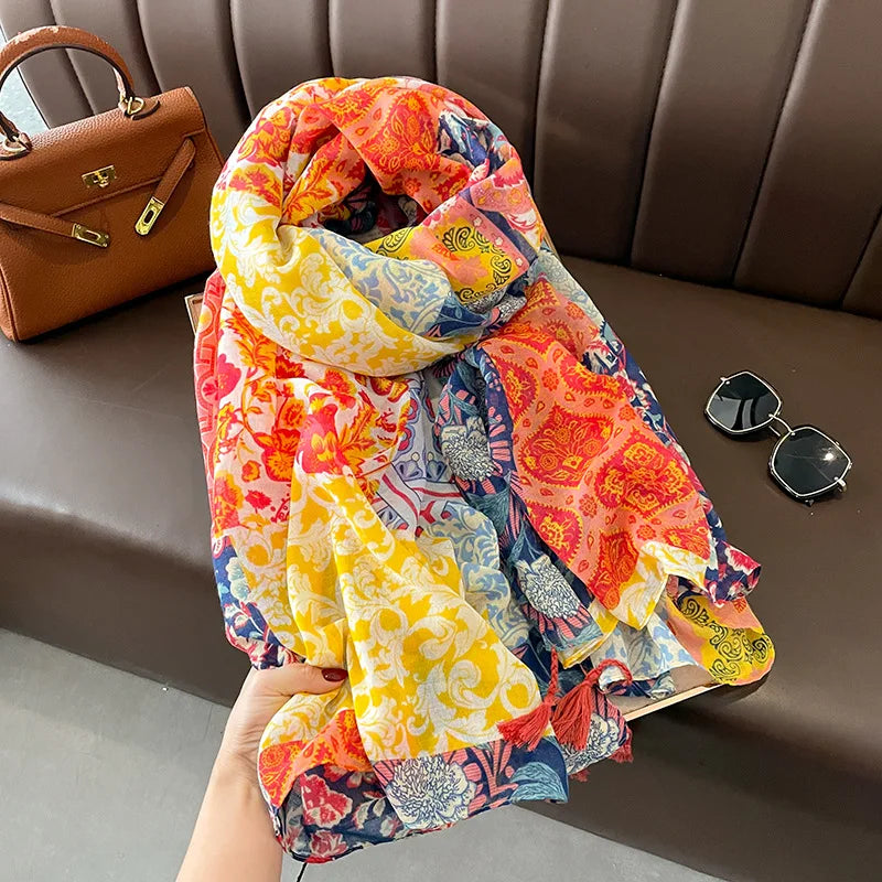 Vibrant floral and paisley-patterned scarf in a mix of yellow, red, blue, and pink hues. The scarf features soft, flowing fabric with tassels at the edges and is accompanied by a brown handbag and gold framed sunglasses placed nearby.