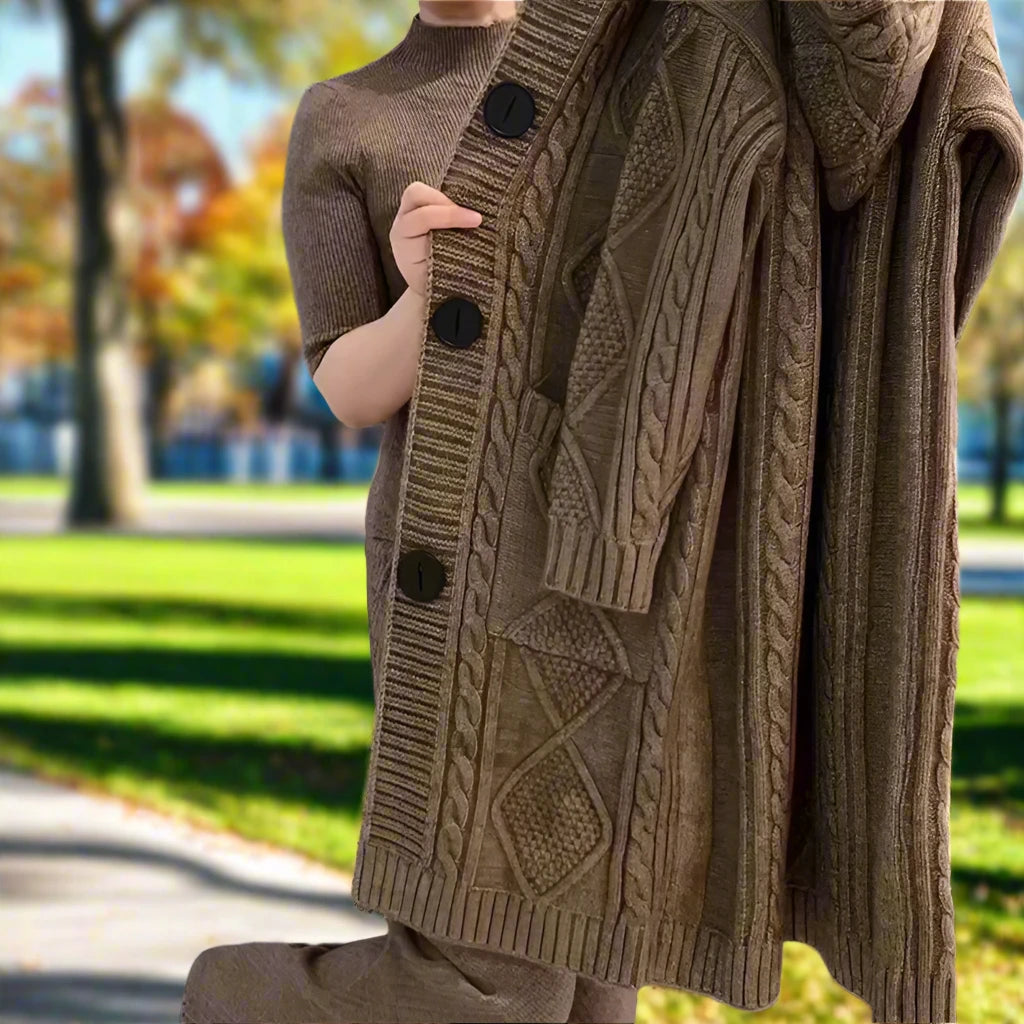  A person is standing in a park with autumn foliage visible in the background. They are holding a textured coffee brown sweater with large buttons and a patterned design, along with matching pants. The person’s face is not visible, and the image captures only from the neck down. The clothing suggests a cozy autumn fashion style, and the setting indicates a crisp outdoor environment.