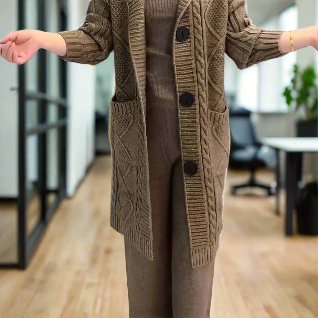 A person stands indoors with arms outstretched, wearing a coffee brown textured cardigan with buttons over a matching set of pants and top. The background is blurred but appears to be an office or room with furniture. The focus is on the cozy, coordinated outfit.