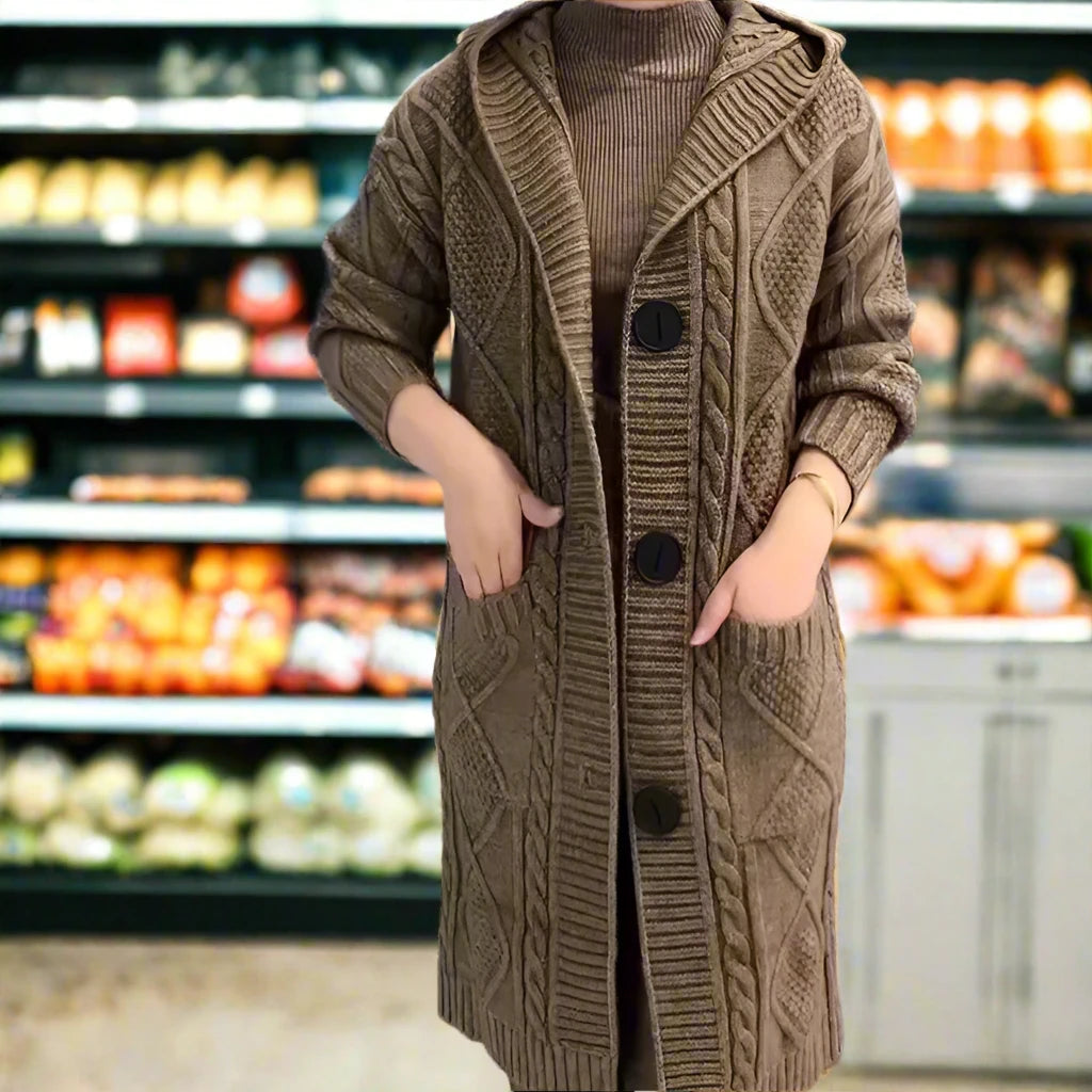 An individual stands in a grocery store aisle, facing the camera without their head visible in the frame. They are wearing a long, coffee brown, jacquard twist cardigan with a hood and large buttons down the front. Hands are placed casually in pockets, and behind the individual is a blurred background featuring shelves stocked with various colorful produce.