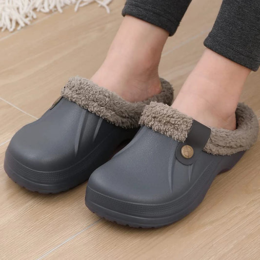 A pair of feet wearing gray clogs with a fuzzy, light brown lining. The clogs have a button detail on the side and are being worn indoors on a wooden floor. The person is wearing dark gray leggings.