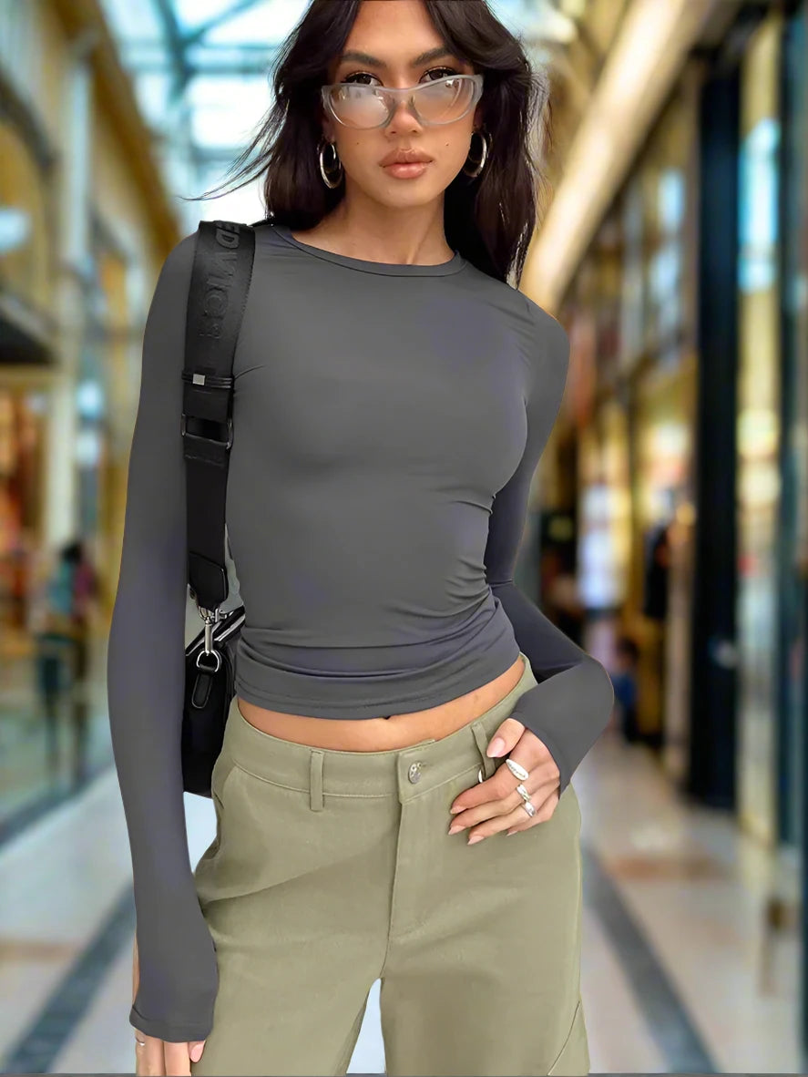 The image shows a young woman standing in a casual pose. She is wearing a light grey, long-sleeve top paired with high-waisted, light colored blue jeans. Her outfit is simple yet stylish, embodying a relaxed and comfortable look. The background is a blurred-out mall scene, keeping the focus on her outfit.