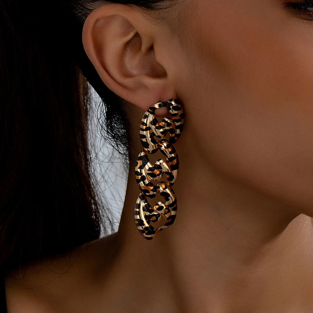 A close-up image showing a person wearing a statement earring. The earring has a bold, multi-link chain design with a leopard print pattern, combining black and gold colors for a striking effect. The earring dangles elegantly, enhancing the person's sophisticated look.