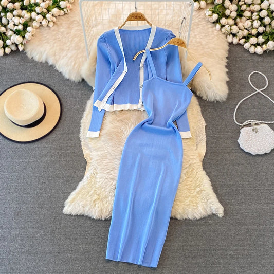 A light blue, two-piece women’s outfit laid out on a white furry surface. The outfit includes a sleeveless, mid-length dress with a cut-out detail on the torso and a matching short-sleeved cardigan with tie closures. Accessories include a white wide-brimmed hat on the left and a small white handbag on the right, surrounded by small white flowers.