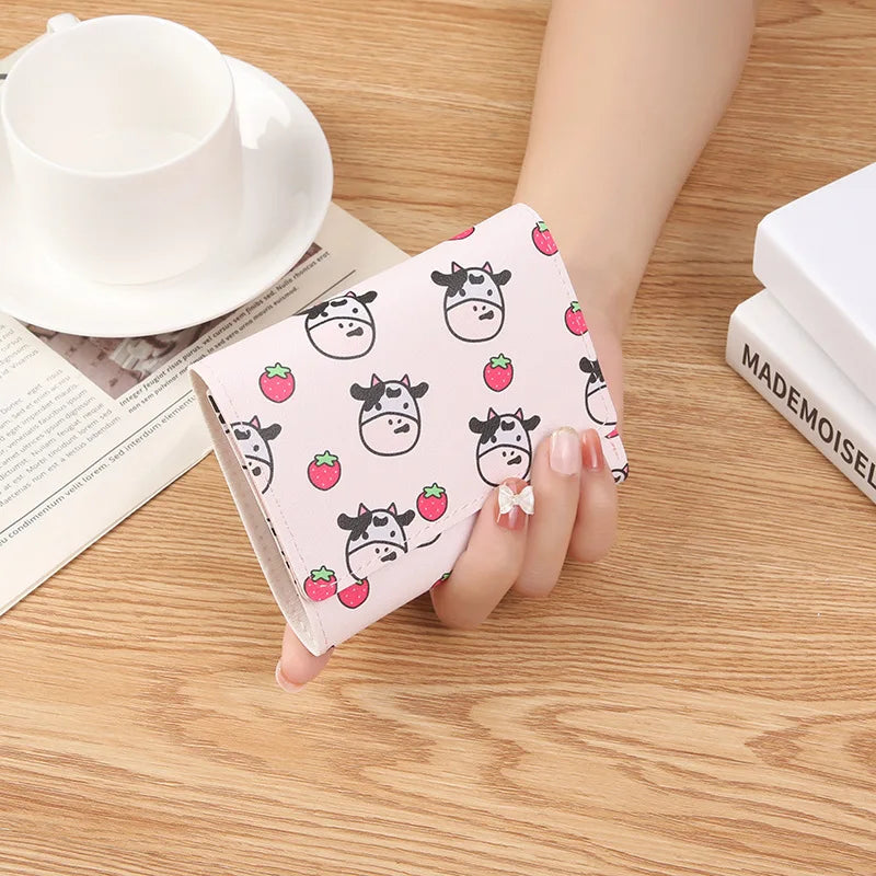 A showcasing of the Light Pink Strawberry Cow Wallet being held in a hand to show a size comparison. This wallet has a cute strawberry and cow print all over on a light pink wallet. 