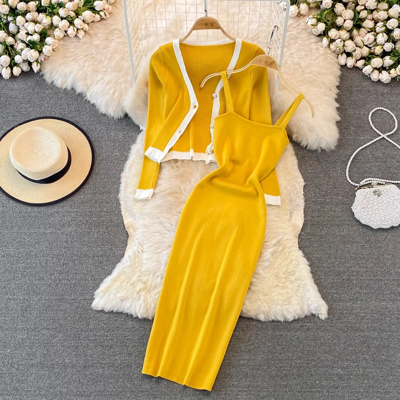 A mustard yellow women’s outfit displayed on a white furry surface. The outfit includes a sleeveless dress with a draped neckline and a matching cardigan with white trim. Accessories include a white handbag and a beige hat on the left side, with white flowers and part of a wicker chair in the background.