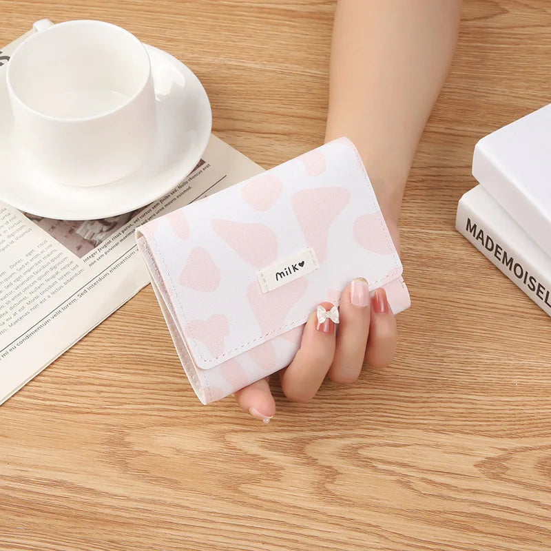 A showcasing of the Pink and White Cow Print Wallet that has a little tag on the front that says milk being held in a hand to show size comparison. The pink is a light shade. 