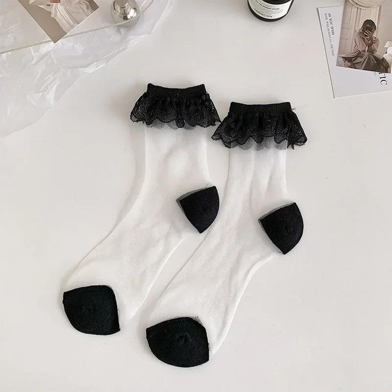 A pair of white, semi-transparent socks with black lace frills at the top are laid out on a white surface. The socks have black reinforced toes and heels. Surrounding the socks are a few items, including a silver spherical object, a bottle, and some photographs.