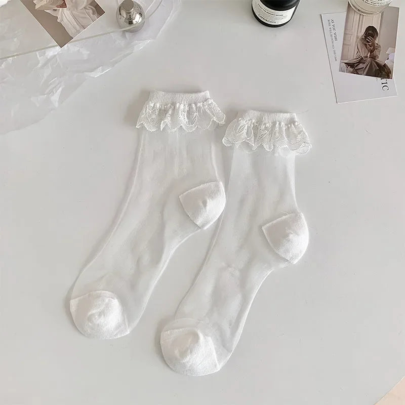 A pair of white, semi-transparent socks with white lace frills at the top are laid out on a white surface. The socks have white reinforced toes and heels. Surrounding the socks are a few items, including a silver spherical object, a bottle, and some photographs.