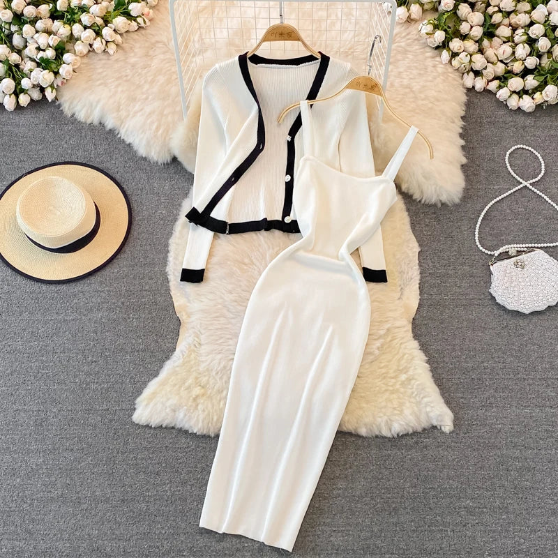 A Pure White women’s outfit displayed on a white furry surface. The outfit includes a sleeveless dress with a draped neckline and a matching cardigan with black trim. Accessories include a white handbag and a beige hat on the left side, with white flowers and part of a wicker chair in the background