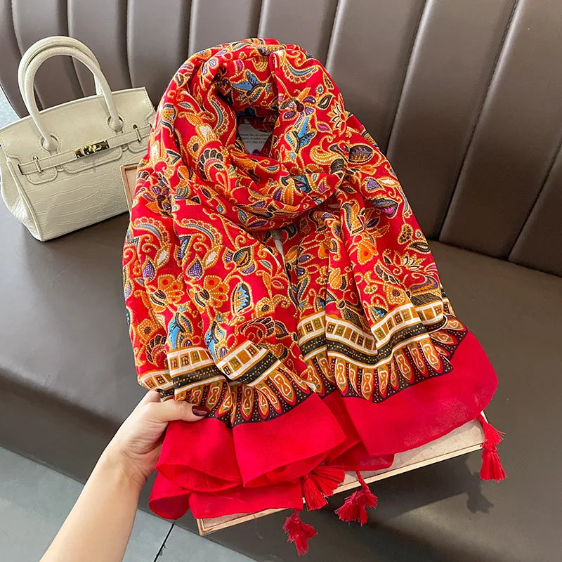 Vibrant red paisley-patterned scarf in a mix of yellow, red, and blue hues. The scarf features soft, flowing fabric with tassels at the edges and is accompanied by a white handbag placed nearby.