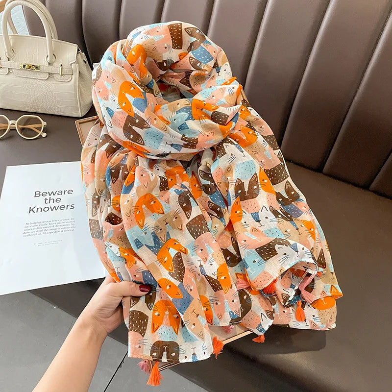 Sheer Cat patterned scarf in a mix of orange, brown, blue, and pink hues. The scarf features soft, flowing fabric with tassels at the edges and is accompanied by a white handbag and white and gold octagonal framed sunglasses placed nearby.