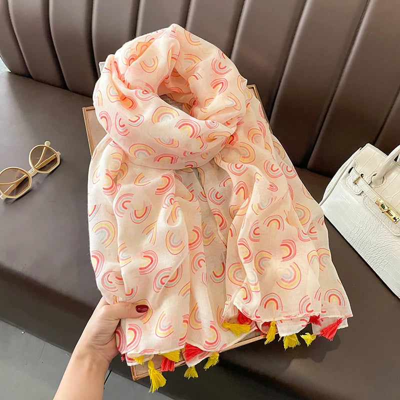 Delicate rainbow patterned sheer scarf in a mix of yellow, red, and pink hues. The scarf features soft, flowing fabric with tassels at the edges and is accompanied by a white handbag and white and gold octagonal framed sunglasses placed nearby.