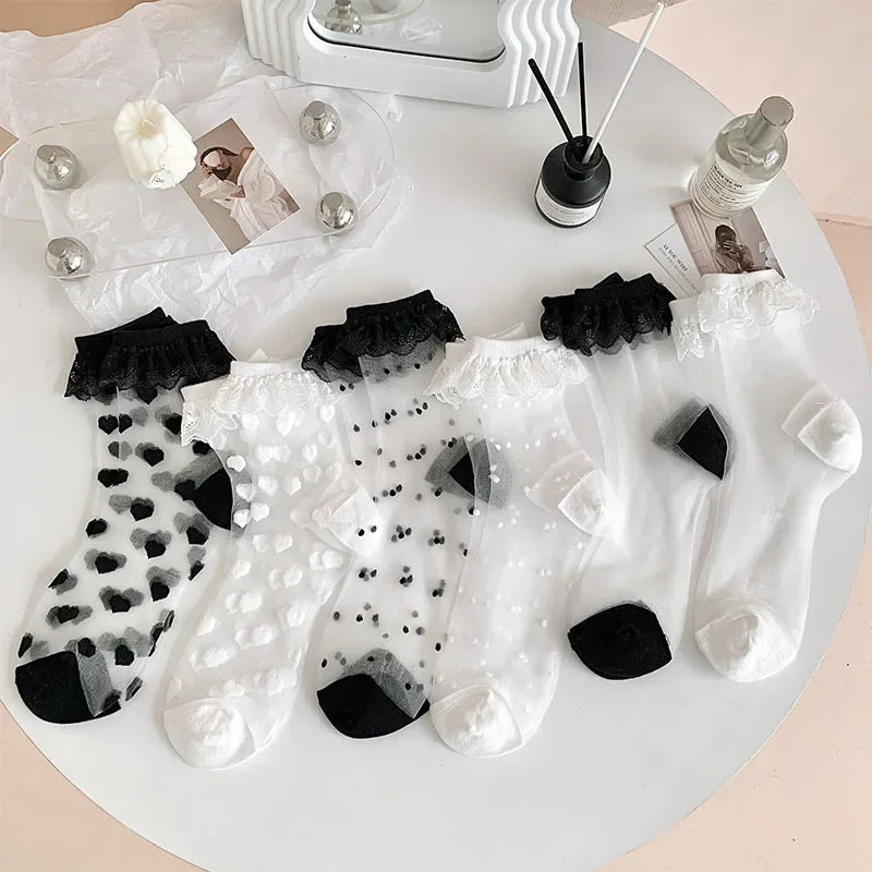 A round white table displays five pairs of sheer socks with different patterns and black toe and heel sections. The socks feature designs such as black hearts, white hearts, black dots, and white dots. The table also holds a decorative tray with a photo, a white flower, and four metallic spheres, along with a black reed diffuser and a clear bottle.