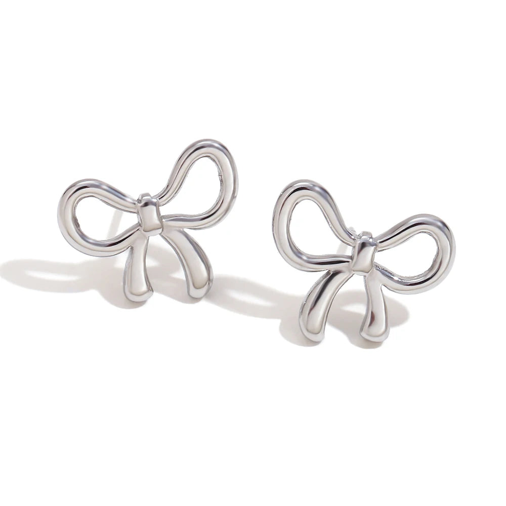 A pair of silver Coquette bow-shaped earrings with a polished finish. The earrings are designed to resemble delicate bows, with loops and tails, and are placed on a white background. The shadows cast by the earrings are visible, adding depth to the image.