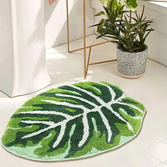 An oval-shaped rug with a white background featuring a detailed tropical monstera leaf pattern in various shades of green. The leaf design includes prominent veins and sections, giving the rug a natural and vibrant appearance staged in bathroom.