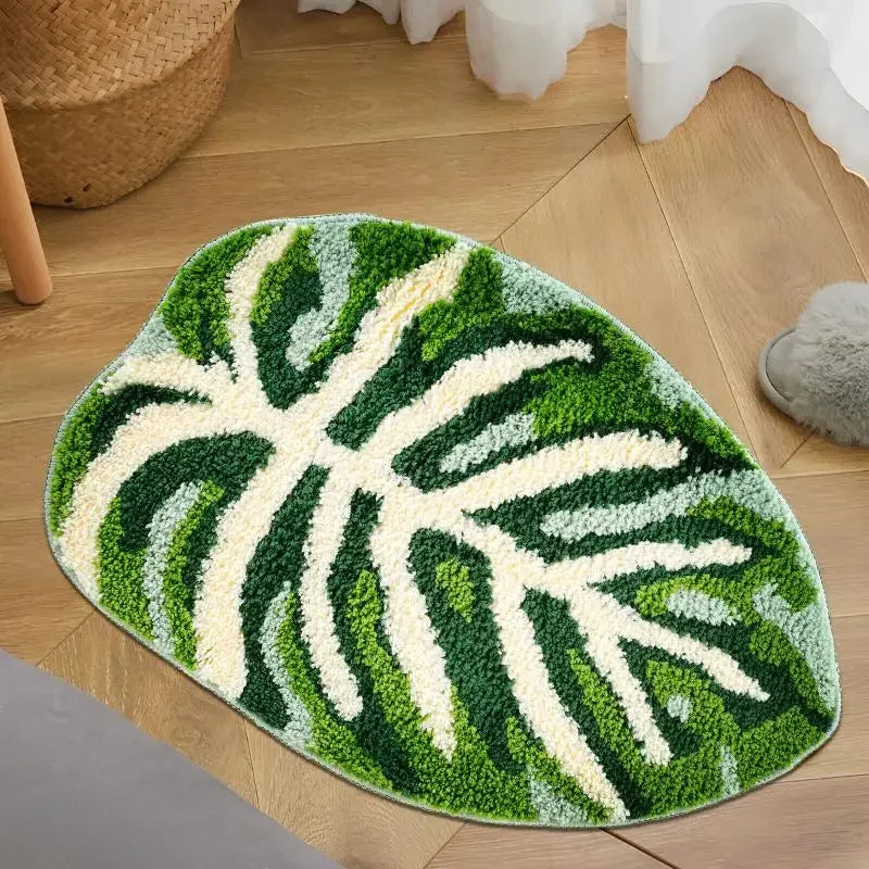 An oval-shaped rug with a brown background featuring a detailed tropical monstera leaf pattern in various shades of green. The leaf design includes prominent veins and sections, giving the rug a natural and vibrant appearance staged in a bathroom.