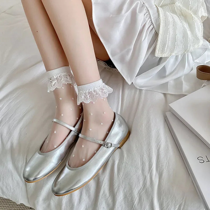 The image shows a close-up of a person's legs and feet. The person is wearing sheer white socks with white polka dots and white lace trim, paired with shiny silver Mary Jane shoes. The person is sitting on a white bedspread, and part of a white dress is visible. There are also some books placed on the bed next to the person.