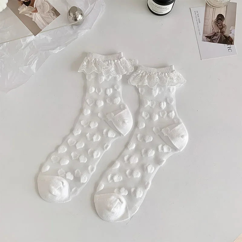 A pair of white, semi-transparent socks with a white heart pattern and white lace frills at the top are laid out on a white surface. The socks have reinforced white toes and heels. Surrounding the socks are a few items, including a silver spherical object, a bottle, and some photographs.