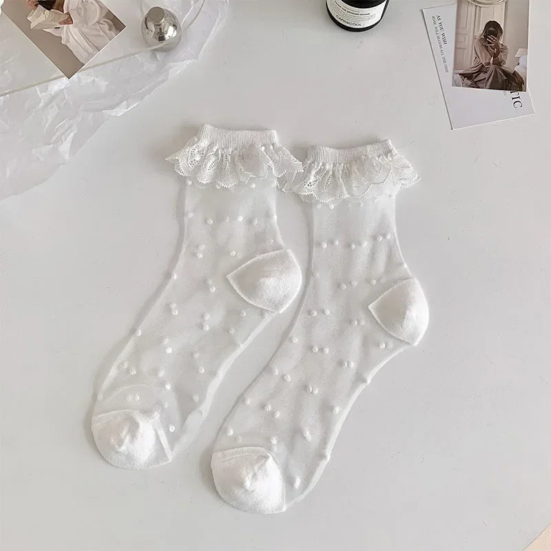 A pair of white, semi-transparent socks with a white dot pattern and white lace frills at the top are laid out on a white surface. The socks have white reinforced toes and heels. Surrounding the socks are a few items, including a silver spherical object, a bottle, and some photographs.