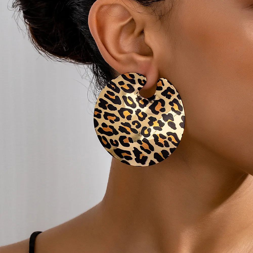 A pair of round earrings with a leopard print pattern. The earrings are flat and have a small cut-out near the top for the earring hook. They are being worn on the ear of a woman to show what it looks like worn. 